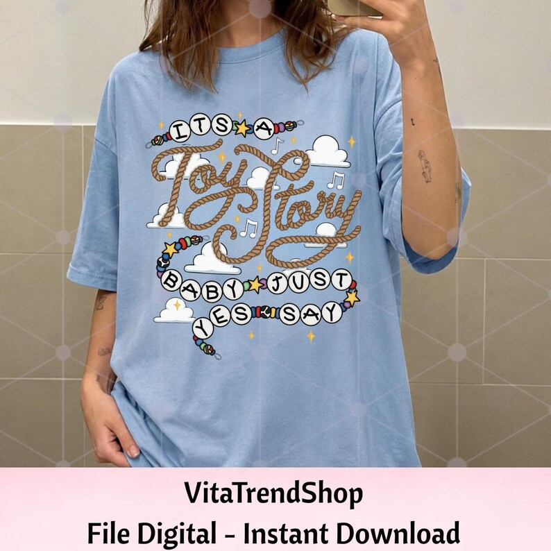 May include: A light blue t-shirt featuring a graphic print with the text "ITS A Toy Story BABY JUST YES SAY". The design includes brown rope-style lettering, white clouds, musical notes, yellow stars, and a string of colorful beads.