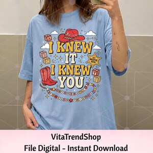 Pu&ograve; includere: Una t-shirt azzurra con una stampa grafica che riporta il testo I KNEW IT I KNEW YOU in giallo. Il design include un cappello da cowboy rosso, stivali da cowboy rossi, cubi dell'alfabeto, una stella da sceriffo BESTIE 4EVA e una collana.