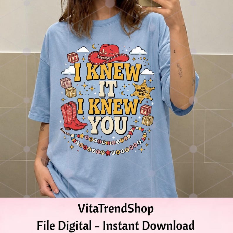 Pu&ograve; includere: Una t-shirt azzurra con una stampa grafica che riporta il testo I KNEW IT I KNEW YOU in giallo. Il design include un cappello da cowboy rosso, stivali da cowboy rossi, cubi dell'alfabeto, una stella da sceriffo BESTIE 4EVA e una collana.