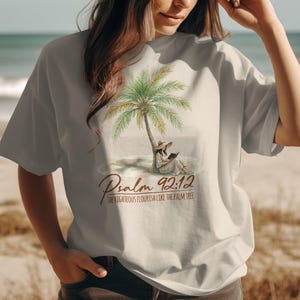 May include: A light gray t-shirt featuring a watercolor illustration of a palm tree on a beach. The design includes a person reading under the tree and the text "Psalm 92:12".
