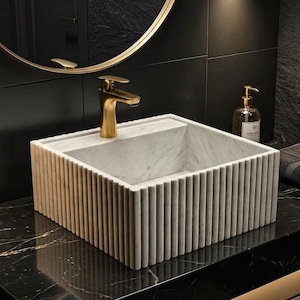 Puede incluir: Un lavabo rectangular de mármol estriado con un grifo dorado. El lavabo está colocado contra una pared de azulejos negros con un espejo con marco dorado. También se ven un dispensador de jabón y una toalla doblada.