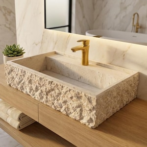 Peut inclure: Meuble-lavabo de salle de bain moderne avec un évier rectangulaire en pierre et un robinet doré. L'évier repose sur un meuble en bois clair avec une étagère inférieure contenant des serviettes pliées. Une plante succulente dans un pot blanc est sur le comptoir.