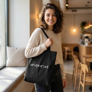 May include: A black tote bag with the coordinates 48°45'N 8°14'E printed in white. The bag is held by a person wearing a cream-colored sweater and dark jeans. The background shows a window and a cafe setting.