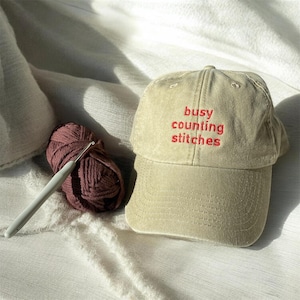 Könnte beinhalten: Beige Baseball Cap mit dem roten gestickten Text "busy counting stitches". Ein Knäuel bordeauxfarbener Wolle und eine Häkelnadel liegen neben der Kappe. Die Artikel befinden sich auf einer weißen, strukturierten Oberfläche.