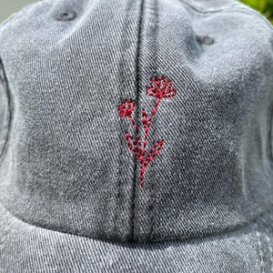 Puede incluir: Gorra de béisbol de mezclilla gris con un diseño floral rojo bordado en la parte delantera. La gorra tiene una visera curva y un cierre de correa de tela. El bordado floral añade un toque de color al sombrero casual.