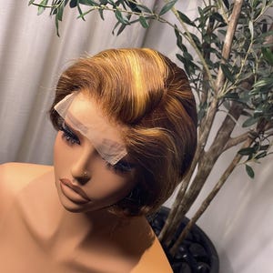 May include: A mannequin head displaying a short, brown wig with blonde highlights. The wig features a lace front and is styled with soft waves. The mannequin is positioned in front of a potted plant with green leaves.