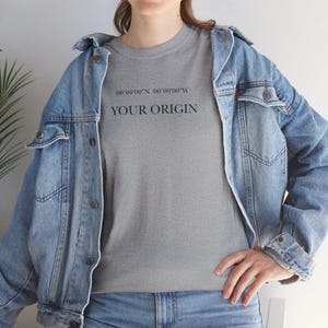 Könnte beinhalten: Ein graues T-Shirt mit dem Text "YOUR ORIGIN" und Koordinaten in Schwarz gedruckt. Die Person trägt eine hellblaue Jeansjacke. Das T-Shirt hat einen Rundhalsausschnitt.