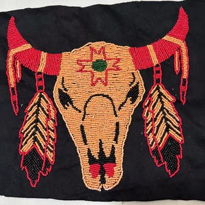 May include: A black fabric square featuring a beaded design of a bull skull. The skull is primarily orange, with red and black accents. The horns are red and orange, and feathers hang below. A green star is in the center of the skull.