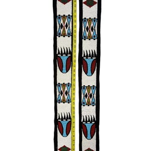 May include: Two long, narrow strips of black fabric featuring white beaded panels with geometric patterns in red, blue, and green. A yellow measuring tape is placed along the length, showing the strips measure approximately 38 inches long.