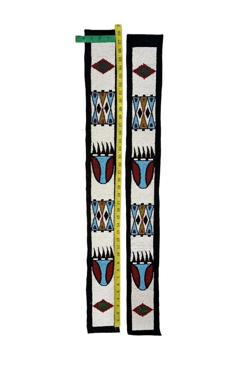 May include: Two long, narrow strips of black fabric featuring white beaded panels with geometric patterns in red, blue, and green. A yellow measuring tape is placed along the length, showing the strips measure approximately 38 inches long.