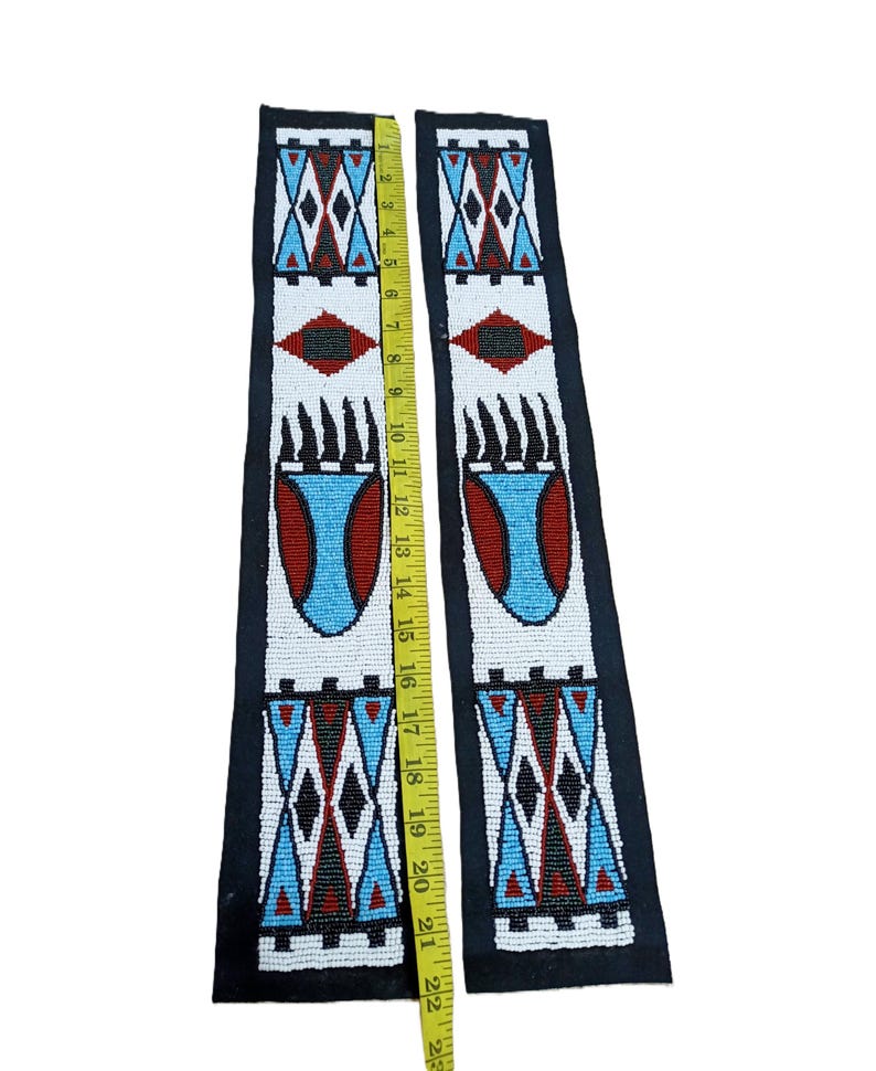 May include: Two long, rectangular strips of black fabric decorated with intricate white, red, black, and light blue beadwork in geometric and bear claw patterns. A yellow measuring tape placed between the strips shows a length of 22 inches.