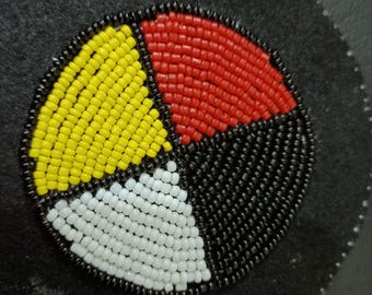May include: A circular patch featuring a four-quadrant design created with small glass beads. The quadrants are colored in yellow, red, white, and black, separated by thin lines of black beads. The entire beaded design is mounted on a black felt base.
