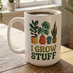 May include: White ceramic mug filled with coffee, featuring a design of potted plants and the text "I GROW STUFF" in green. The mug has a large handle and is set on a wooden surface.