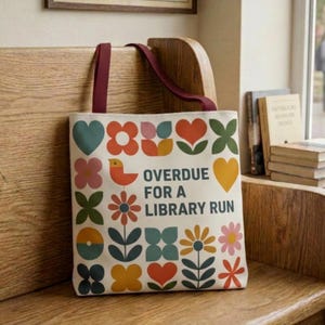 May include: Cream-colored tote bag with a colorful retro design of hearts, flowers, and geometric shapes. The bag features a burgundy strap and the text "OVERDUE FOR A LIBRARY RUN". The bag is sitting on a wooden bench.
