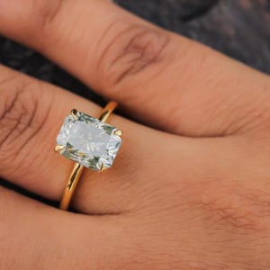 Puede incluir: Un anillo de oro con una gran gema rectangular de color verde claro. La banda del anillo es de oro fino y pulido, y la gema está engastada con cuatro garras. El anillo se muestra en un dedo.