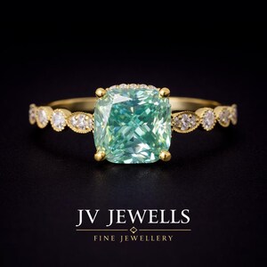 Puede incluir: Un anillo de oro con una gran piedra central de color verde menta en talla cojín sujeta por cuatro garras. La banda está adornada con una serie de pequeñas piedras claras redondas en monturas de oro en forma de marquesa. Texto JV JEWELLS FINE JEWELLERY.