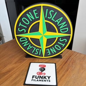 May include: A circular Stone Island logo in black, yellow, and green, displayed on a wooden surface. The logo features the words "STONE ISLAND". A small square with "FUNKY FILAMENTS" is below the logo.