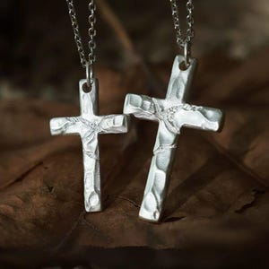 Puede incluir: Dos colgantes de cruz en tono plateado que cuelgan de cadenas de metal a juego. Cada cruz presenta una superficie martillada y texturizada con hendiduras irregulares y un acabado mate, creando una apariencia rugosa en el metal.