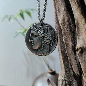 Può includere: Collana con pendente argentato raffigurante un profilo classico su una moneta rotonda. Il pendente è appeso a una catena. Lo sfondo sfocato include elementi naturali come legno e vegetazione.