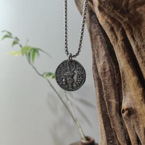 Puede incluir: Un collar con colgante plateado con un dije circular en forma de moneda. El dije tiene un diseño en relieve con una imagen central en forma de antorcha y símbolos circundantes. La cadena del collar es de diseño de eslabones. El fondo incluye madera y vegetación.