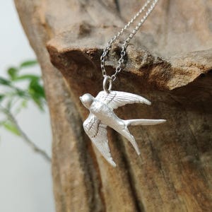 Op de afbeelding: Een zilverkleurige ketting met een hanger in de vorm van een vogel in vlucht met gespreide vleugels en een gevorkte staart. De hanger heeft een matte, getextureerde afwerking en hangt aan een fijne zilverkleurige metalen schakelketting.
