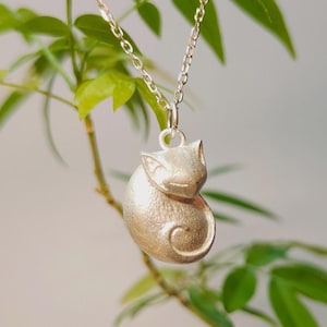 Può includere: Un ciondolo color argento a forma di gatto raggomitolato con superficie testurizzata, sospeso a una catenina in metallo color argento abbinata. La figura del gatto presenta orecchie a punta e una coda che si curva attorno al corpo, con un anellino.