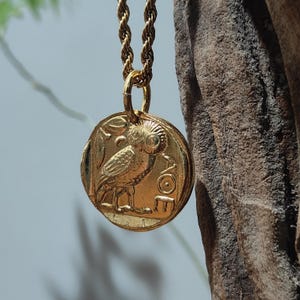 Peut inclure: Collier pendentif doré avec un motif de hibou détaillé sur une pièce de monnaie circulaire. Le hibou est représenté de profil, avec des détails de plumes complexes. Le pendentif est suspendu à une chaîne dorée torsadée, créant un look classique et élégant.