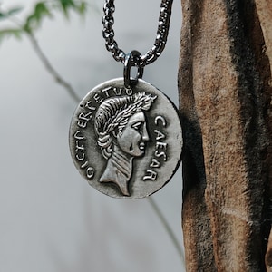 Può includere: Collana con pendente color argento con un design a moneta circolare. La moneta presenta il profilo di una persona con una corona d'alloro e la scritta "CAESAR". Il pendente è appeso a una catena.