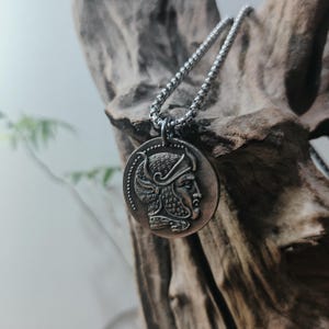 Può includere: Una collana con ciondolo circolare color argento che presenta il profilo in rilievo di una figura che indossa un elmo ornato. Il ciondolo pende da una catena metallica. Il design include un bordo perlato attorno al ciondolo circolare.