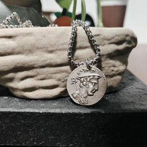 Può includere: Collana argentata con un ciondolo circolare raffigurante un profilo classico. Il ciondolo è appeso a una catena. La collana è mostrata su uno sfondo neutro, evidenziando la sua lucentezza metallica e il design dettagliato.