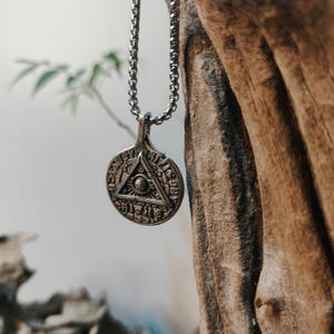 May include: A silver-coloured pendant necklace. The circular pendant features an eye-in-triangle design. The pendant hangs from a chain necklace. The background includes a blurred natural setting with wood and foliage.
