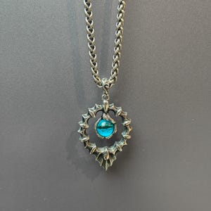 May include: A silver pendant necklace with a circular, decorative design and a central, round, blue gemstone. The pendant is suspended from a thick, textured chain. The necklace is set against a grey background.