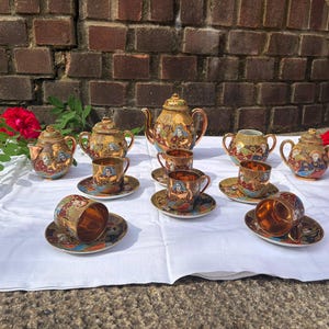 May include: A tea set featuring a teapot, sugar bowl, creamer, and six cups with saucers. Each piece is crafted from dark brown ceramic with a metallic gold finish, decorated with colourful, detailed illustrations of figures in traditional attire.