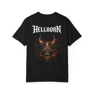 May include: Black t-shirt featuring the word "HELLBORN" in white gothic lettering above a detailed illustration of a demonic face with fiery horns and glowing eyes. The design is centered on the shirt.