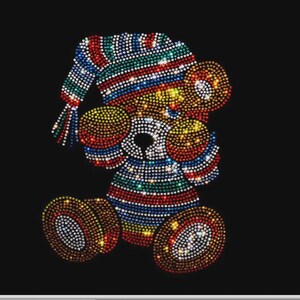Op de afbeelding: Een teddybeerontwerp gemaakt met veelkleurige strass-steentjes op een zwarte achtergrond. De beer draagt een gestreepte slaapmuts en een bijpassend gestreept shirt, met cirkelvormige accenten op de oren, handen en voeten van fonkelende steentjes.