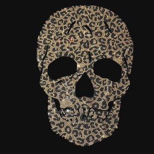 May include: A skull-shaped iron-on patch or transfer design featuring a dense pattern of shimmering gold and black rhinestones arranged in a leopard print motif. The design has hollow eye sockets and a nasal cavity, creating a detailed skull silhouette.