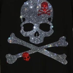 Könnte beinhalten: Eine schwarze Stoffoberfläche mit einem großen Totenkopf- und Knochenmotiv aus klaren, reflektierenden Strasssteinen. Der Totenkopf enthält ein kleineres rotes Strass-Totenkopf- und Knochendetail auf der Stirn sowie einen roten Totenkopf.