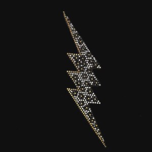 May include: A lightning bolt shaped patch featuring a dense arrangement of small, reflective rhinestones in shades of gold, silver, and black. The design is oriented vertically against a solid black background, creating a high-contrast visual effect.