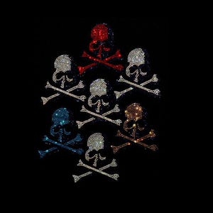May include: A collection of seven skull and crossbones motifs arranged in a cluster against a black background. Each skull is composed of small, sparkling rhinestones in various colors including red, white, blue, orange, and clear crystal.