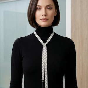 Puede incluir: Un collar tipo lazo largo de tono plateado con una banda de malla plana y flexible que presenta un patrón geométrico de chevrón. El collar termina en dos cadenas plateadas colgantes con pequeñas cuentas esféricas en las puntas, sobre un cuello alto negro.