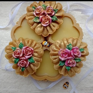 May include: A decorative, floral-themed item with a light yellow base and three gold-colored, flower-shaped embellishments. Each flower features pink roses and green leaves. Small bee-shaped accents are also present, along with white ribbons.