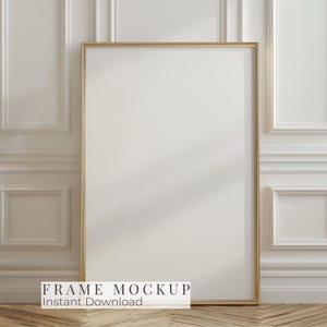 Puede incluir: Una maqueta de marco rectangular con marco dorado apoyada contra una pared blanca con molduras. El marco está vacío, con el texto "FRAME MOCKUP Instant Download" en la parte inferior. El suelo es de madera.