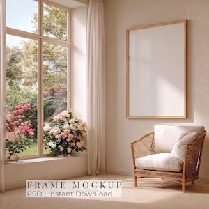 May include: A vertical rectangular wooden frame with a blank white centre hangs on a beige wall next to a wicker armchair with white cushions. A large window with white curtains reveals a garden with pink flowers and green foliage outside.