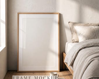 May include: A vertical wooden picture frame with a white mat and a blank rectangular center, positioned on a wooden floor next to a small white ceramic mug and a stack of two books, with the text FRAME MOCKUP PSD Instant Download.