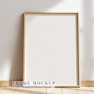 May include: A vertical rectangular light wood picture frame with a blank white center. The frame is positioned upright. The text "FRAME MOCKUP PSD Instant Download" is printed in black at the bottom center of the image area.