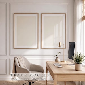 May include: Two vertical rectangular wooden frames with white matting and blank cream-colored inserts mounted on a white paneled wall above a wooden desk featuring a computer monitor, a small potted plant, and a gold desk lamp.