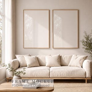 May include: Two vertical rectangular frames with light wood borders mounted on a beige wall above a beige sofa. The sofa features several matching beige cushions. A small potted plant sits on a wooden table in the foreground.