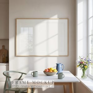 May include: A horizontal rectangular picture frame with a light wood finish, featuring a white mat board inside. The frame is mounted on a plain wall above a white table, which holds a bowl of fruit, a blue pitcher, and two green mugs.