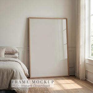 Puede incluir: Una maqueta de marco rectangular de madera, grande, apoyada contra una pared en un dormitorio. El marco está vacío, con el texto "FRAME MOCKUP PSD - Instant Download" en la parte inferior. La habitación cuenta con una cama con ropa de cama de tonos neutros y una ventana con cortinas transparentes.