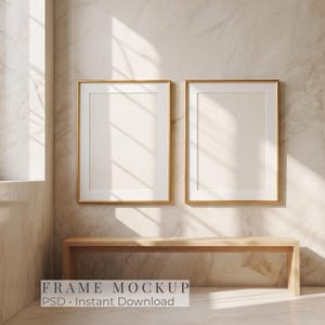 May include: Two vertical rectangular picture frames with thin gold borders and white matting, mounted side by side on a light beige marble wall above a simple wooden bench. The scene features soft natural light casting window pane shadows.